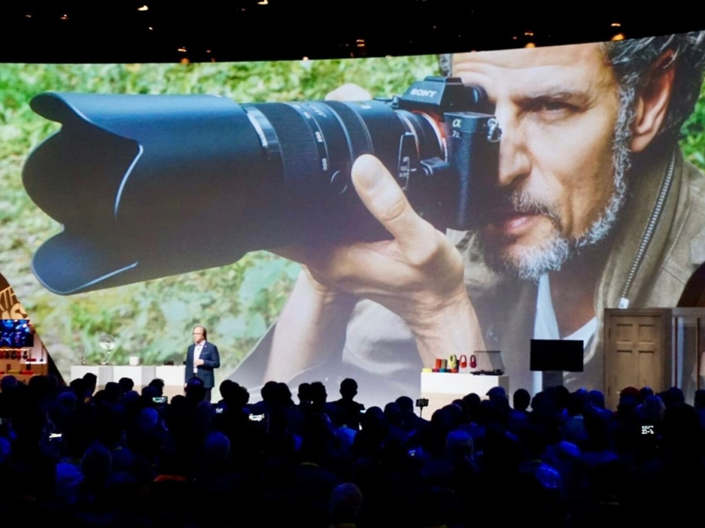 Audience watches a large screen showing a man with a camera and telephoto lens.