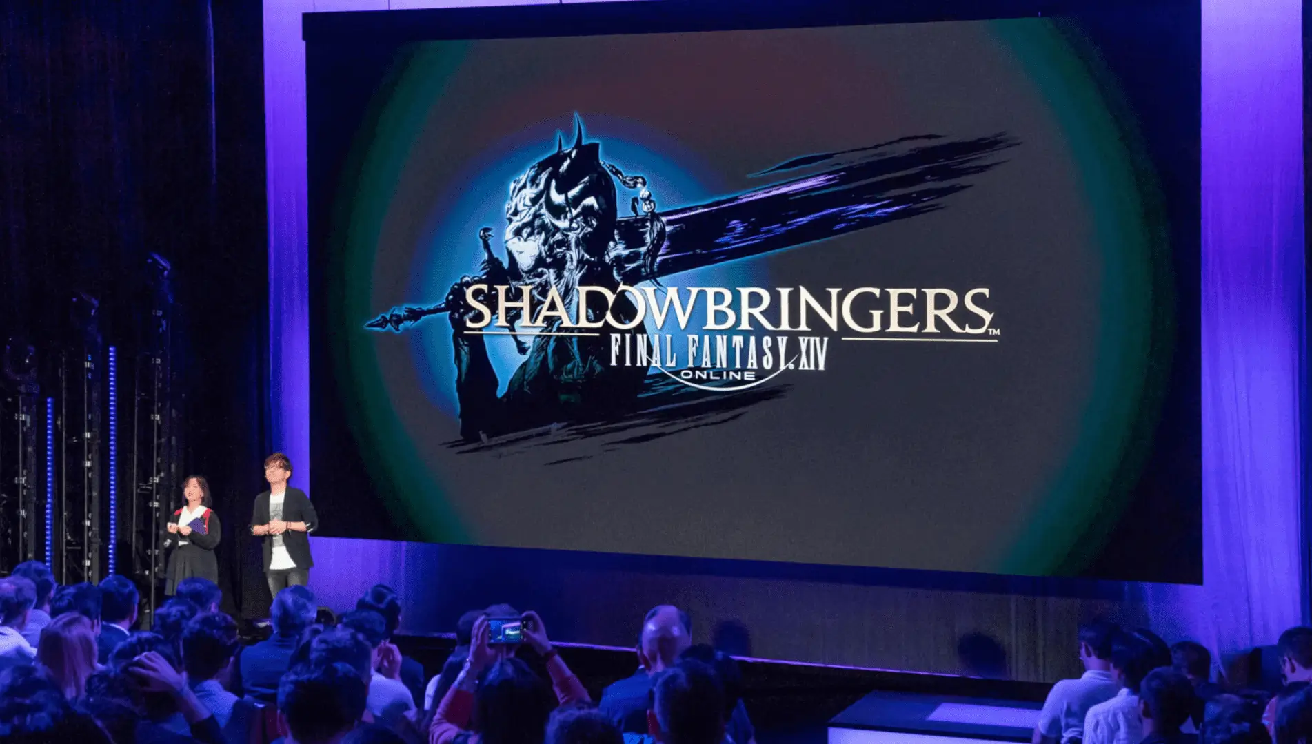 Two hosts on stage beside Shadowbringers display.